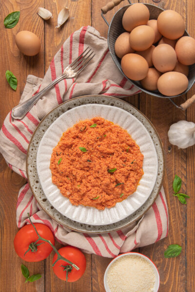 Uova strapazzate al pomodoro su canovaccio bianco a righe rosse, cestino di uova, pomodoro, aglio e foglioline di basilico