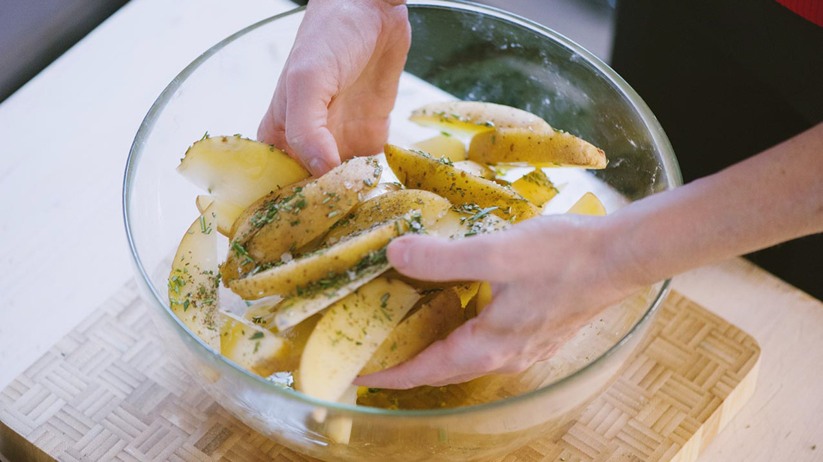 Mani che mescolano spicchi di patate con erbe aromatiche e olio in una ciotola