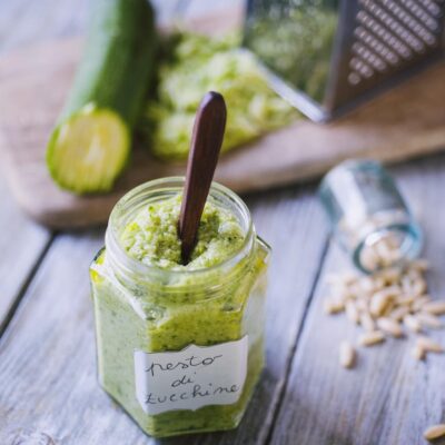 Pesto di zucchine in vasetto con etichetta scritta a mano, cucchiaino immerso, foglie di basilico e pinoli intorno