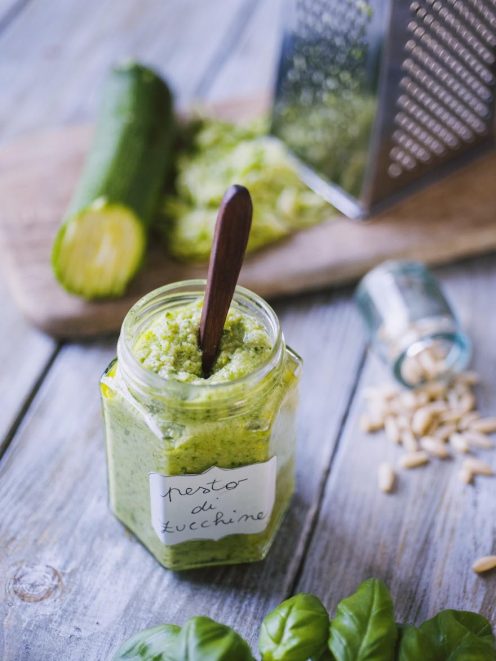 Pesto di zucchine in vasetto con etichetta scritta a mano, cucchiaino immerso, foglie di basilico e pinoli intorno