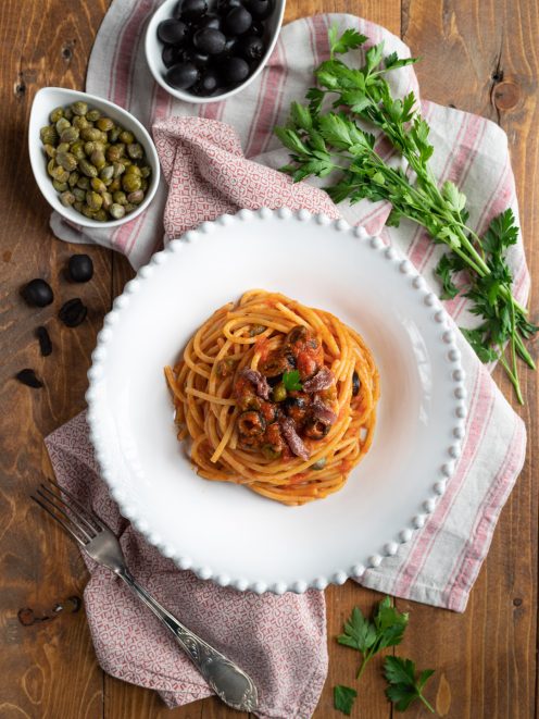 Spaghetti al sugo alla puttanesca con olive nere, capperi e acciughe, guarniti con prezzemolo fresco