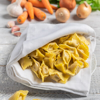 Agnolotti del plin freschi all'uovo adagiati su un canovaccio, con carote, cipolle e aglio sullo sfondo