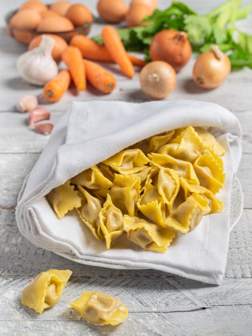 Agnolotti del plin freschi all'uovo adagiati su un canovaccio, con carote, cipolle e aglio sullo sfondo