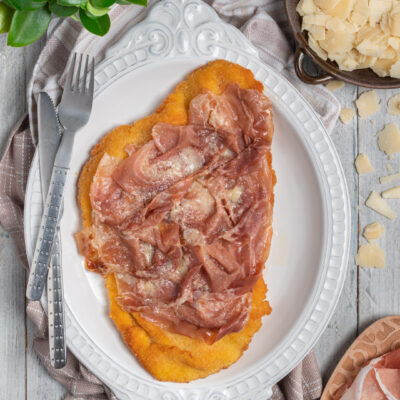 Cotoletta alla bolognese impanata e fritta, coperta di prosciutto crudo e scaglie di formaggio