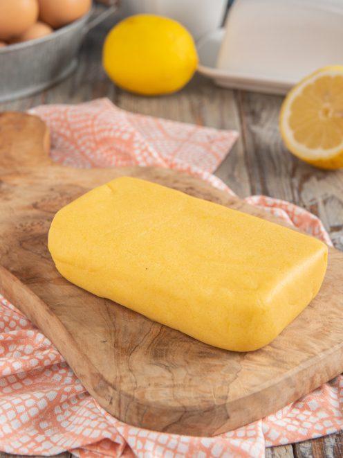 Pasta frolla senza glutine a panetto compatto su tagliere in legno, con limoni sullo sfondo