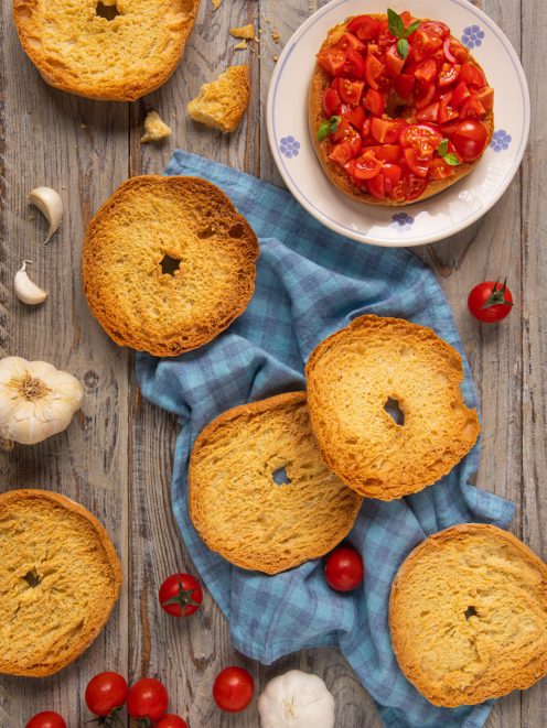 Friselle pugliesi con pomodorini freschi a pezzi e foglie di basilico, accanto ad aglio e pomodorini interi