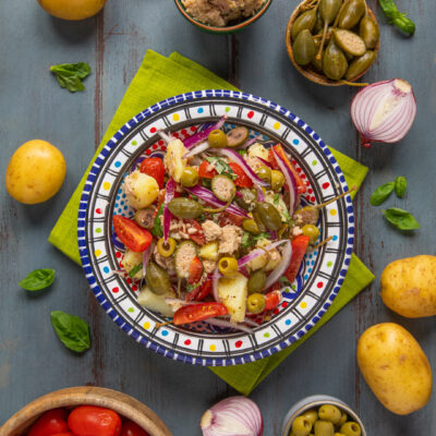 Insalata eoliana con patate, tonno, pomodori, olive, capperi e cipolla rossa circondata dagli ingredienti freschi