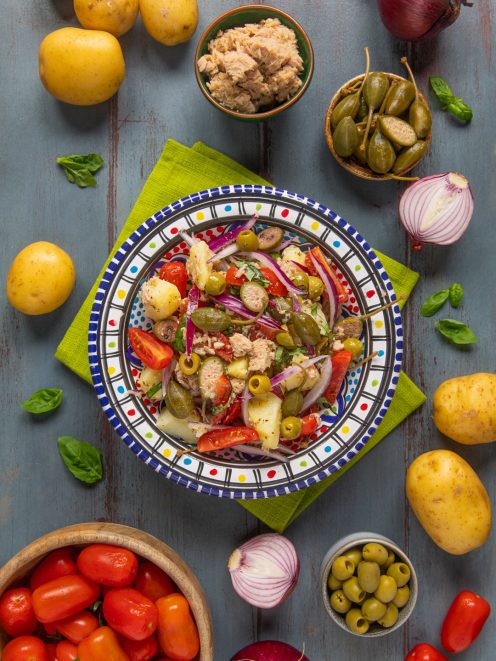 Insalata eoliana con patate, tonno, pomodori, olive, capperi e cipolla rossa circondata dagli ingredienti freschi
