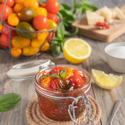 Confettura di pomodorini in vasetto con foglie di basilico, pomodori freschi e limone sullo sfondo