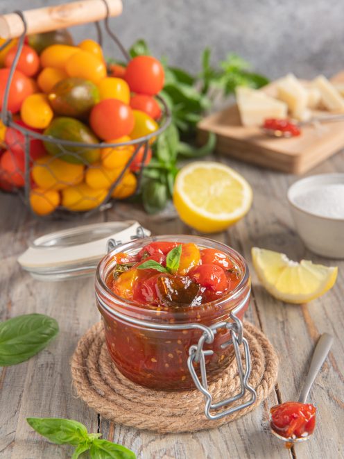 Confettura di pomodorini in vasetto con foglie di basilico, pomodori freschi e limone sullo sfondo