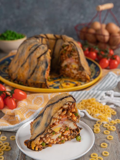 Anelletti alla siciliana in forma a cupola rivestita di melanzane, con una fetta tagliata che rivela il ripieno
