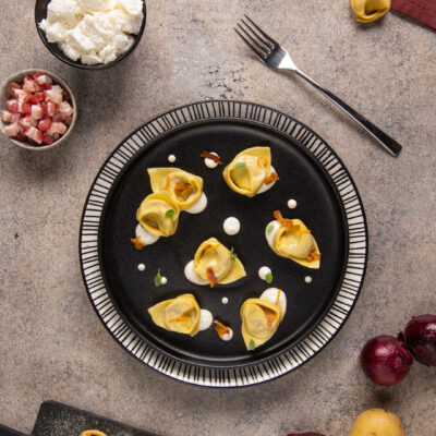 Cappellacci ripieni di cipolla, pancetta e ricotta disposti su piatto scuro con salsa di caprino e pancetta croccante