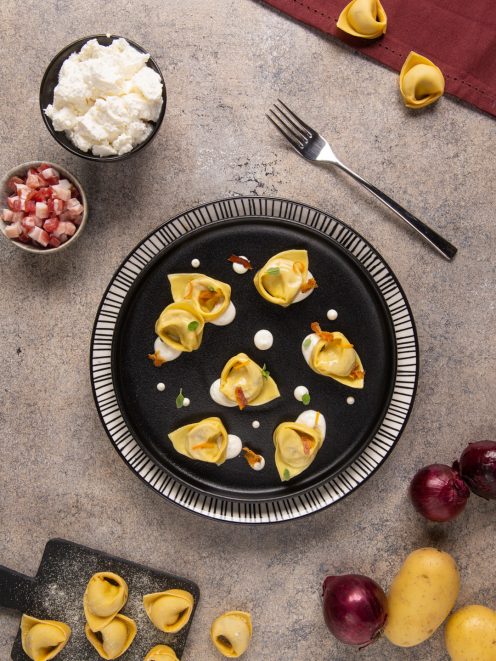 Cappellacci ripieni di cipolla, pancetta e ricotta disposti su piatto scuro con salsa di caprino e pancetta croccante