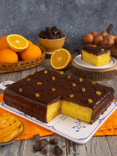 Torta arancia e cioccolato con glassa lucida e cubetti di scorza candita, una fetta tagliata rivela il pan di spagna soffice