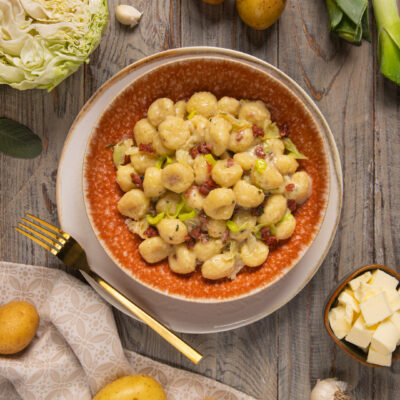 Gnocchi alla crema di verza e cotechino serviti in ciotola circondati da patate, porri e cavolo