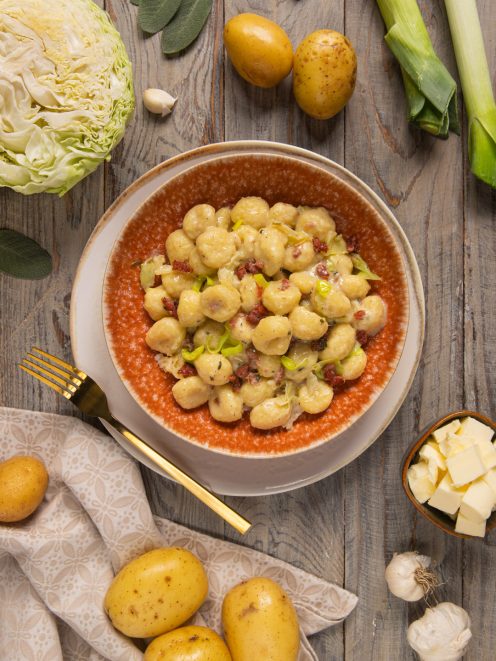Gnocchi alla crema di verza e cotechino serviti in ciotola circondati da patate, porri e cavolo