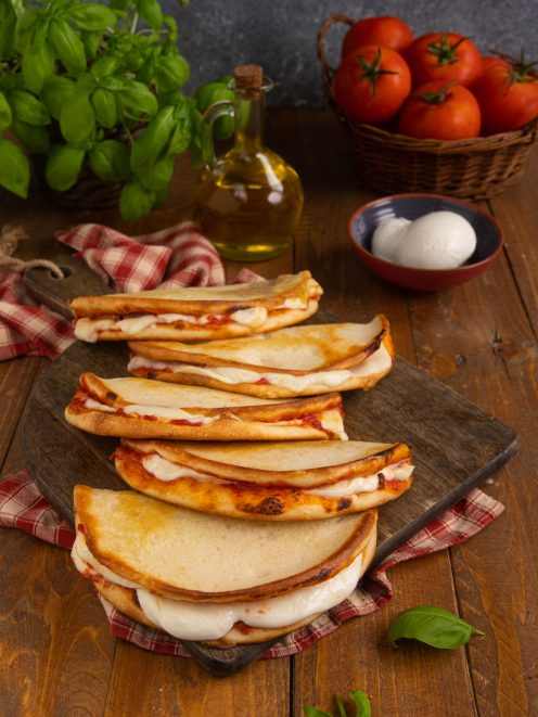 Pizzette da passeggio piegate a mezzaluna con mozzarella filante e pomodoro, disposte in fila su tagliere