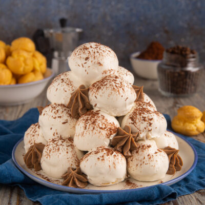 Profiteroles al tiramisù impilati a piramide con glassa bianca, ciuffi di crema al caffè e cacao in polvere