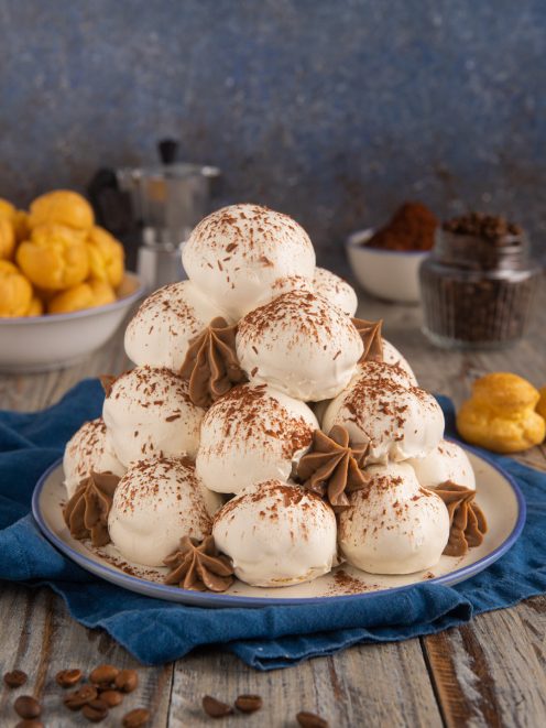 Profiteroles al tiramisù impilati a piramide con glassa bianca, ciuffi di crema al caffè e cacao in polvere