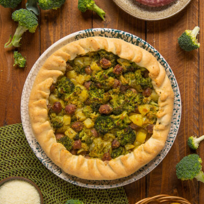 Torta salata con broccoli, patate e salsiccia dal bordo rustico, circondata dagli ingredienti freschi