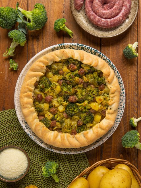Torta salata con broccoli, patate e salsiccia dal bordo rustico, circondata dagli ingredienti freschi