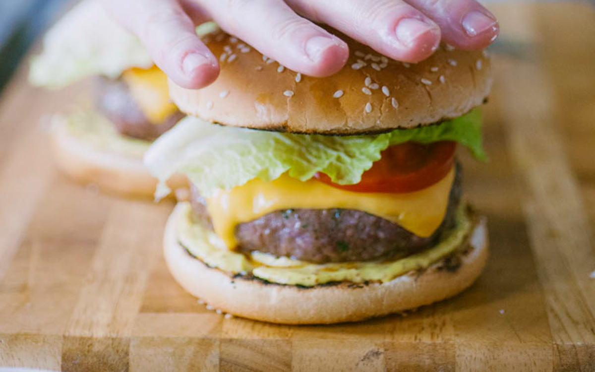 Una mano preme il bun di sesamo sull'hamburger farcito con cheddar fuso, pomodoro e lattuga