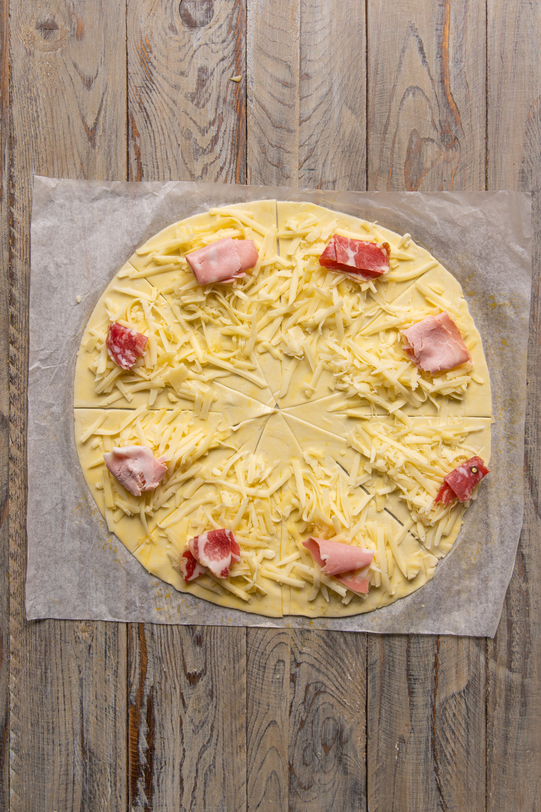 Disco di pasta sfoglia tagliato a spicchi con formaggio grattugiato e pezzetti di salume disposti sul bordo
