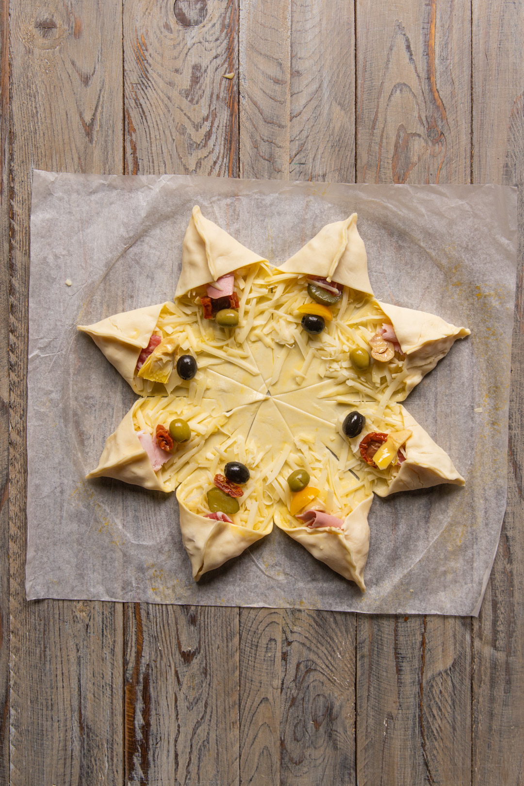 Stella di sfoglia stesa con punte ripiegate su formaggio grattugiato, olive, prosciutto e pomodori secchi
