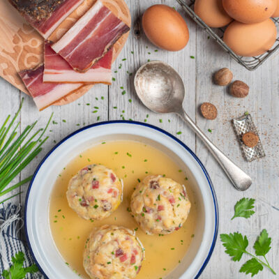Canederli allo speck in brodo dorato con erba cipollina, circondati da uova, speck affettato e noce moscata