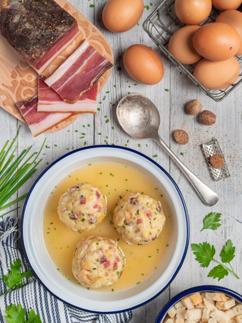 Canederli allo speck in brodo dorato con erba cipollina, circondati da uova, speck affettato e noce moscata