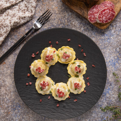 Ravioli con fonduta e briciole di salame disposti a cerchio, guarniti con erbe fresche