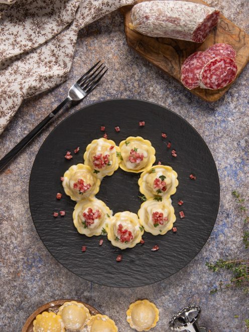 Ravioli con fonduta e briciole di salame disposti a cerchio, guarniti con erbe fresche