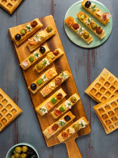 Waffle salati a striscia guarniti con salmone, olive, pomodori secchi e crema su tagliere