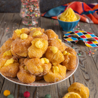 Frittelle di Carnevale ripiene di crema pasticcera, impanate nello zucchero e impilate in abbondanza