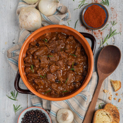 Gulasch triestino in tegame di terracotta con rosmarino e pane tostato accanto a cipolle e paprika