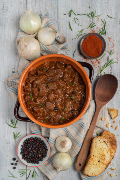 Gulasch triestino in tegame di terracotta con rosmarino e pane tostato accanto a cipolle e paprika