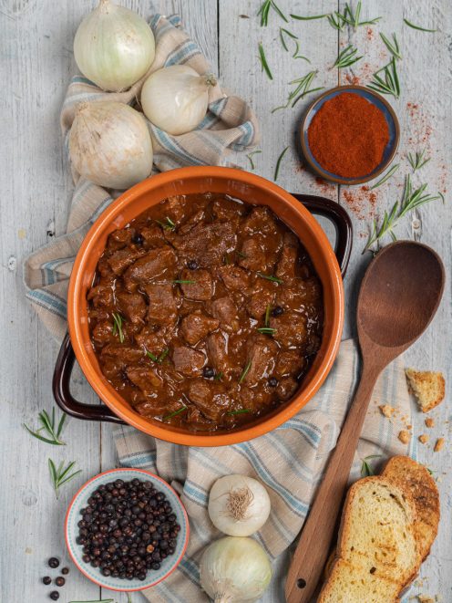 Gulasch triestino in tegame di terracotta con rosmarino e pane tostato accanto a cipolle e paprika