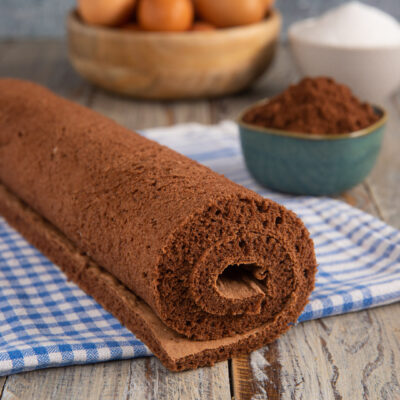 Pasta biscotto al cacao arrotolata su un canovaccio a quadretti con uova e cacao sullo sfondo