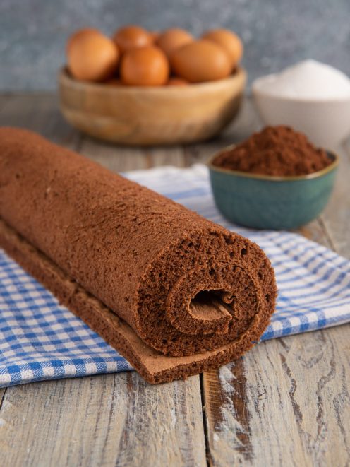 Pasta biscotto al cacao arrotolata su un canovaccio a quadretti con uova e cacao sullo sfondo