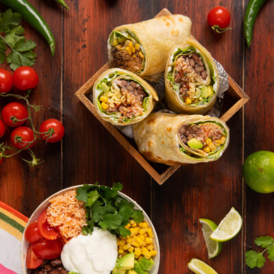 Burrito tagliato a metà con riso, mais, fagioli e avocado, servito con una bowl degli stessi ingredienti