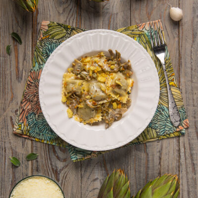 Ravioli di brasato al barolo conditi con carciofi trifolati e granella dorata, circondati da carciofi freschi e aglio