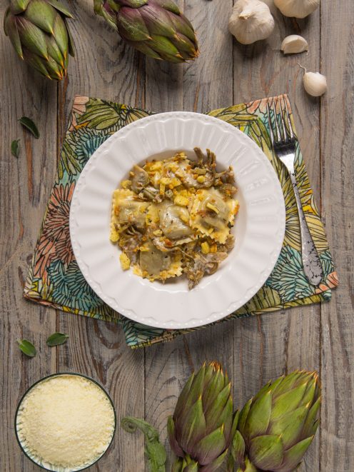 Ravioli di brasato al barolo conditi con carciofi trifolati e granella dorata, circondati da carciofi freschi e aglio