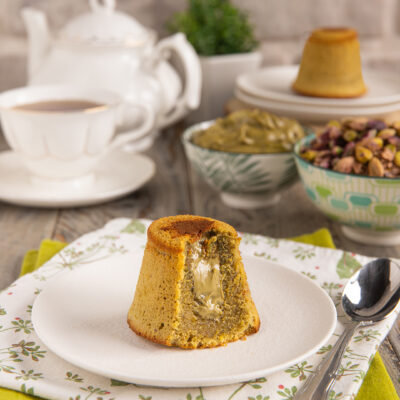 Tortino al pistacchio con cuore fondente tagliato a meta su piattino, pistacchi e crema sullo sfondo