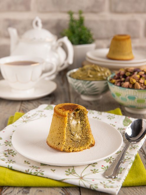 Tortino al pistacchio con cuore fondente tagliato a meta su piattino, pistacchi e crema sullo sfondo