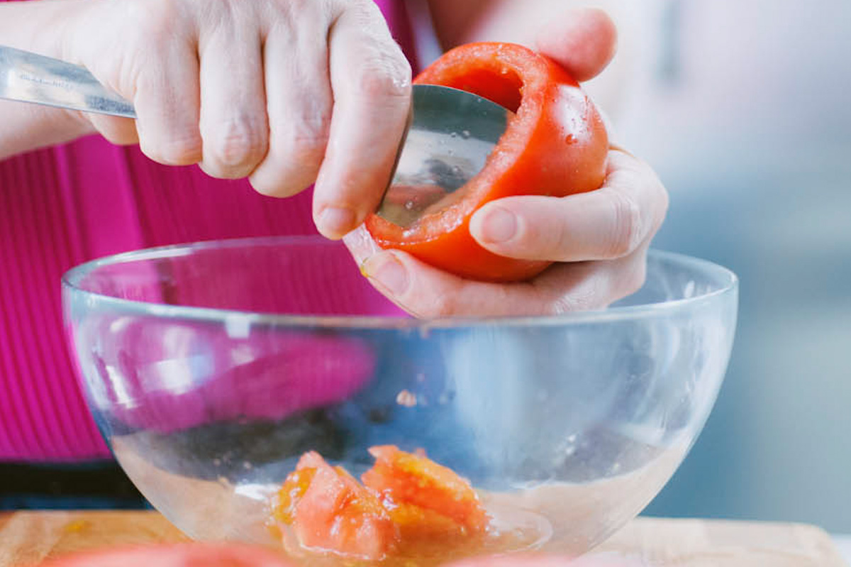 Mani che scavano la polpa di un pomodoro con un cucchiaio raccogliendola in una ciotola