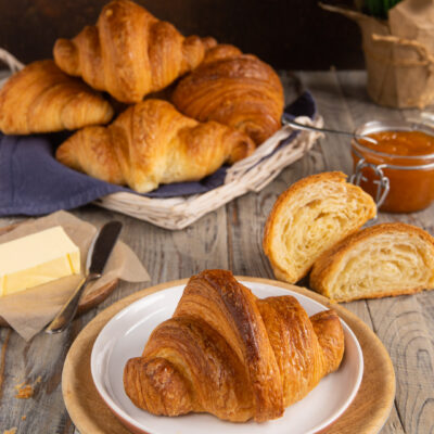 Croissant dorato e sfogliato servito su piatto, con altri croissant, uno tagliato a metà che mostra la mollica soffice