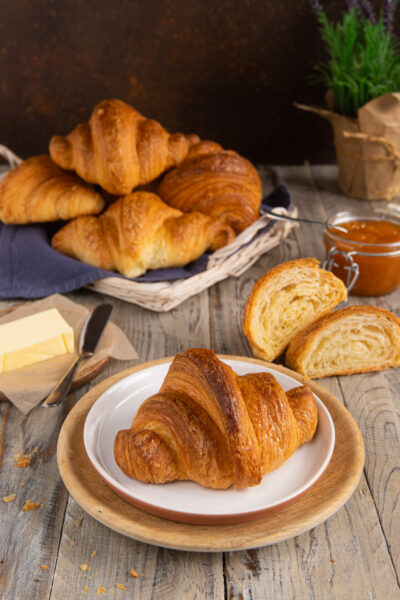 Croissant dorato e sfogliato servito su piatto, con altri croissant, uno tagliato a metà che mostra la mollica soffice