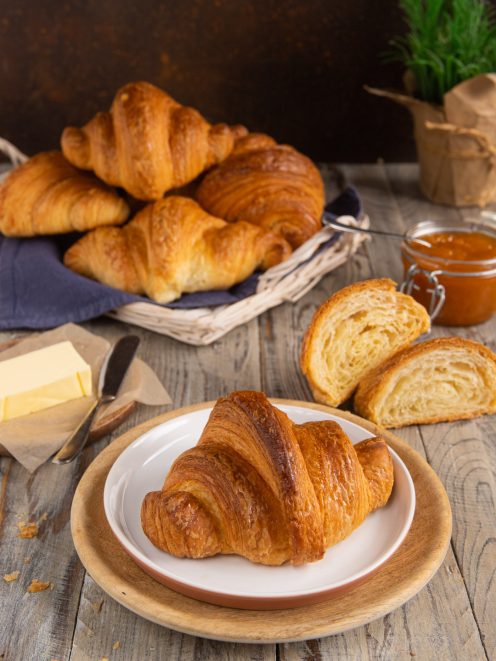 Croissant dorato e sfogliato servito su piatto, con altri croissant, uno tagliato a metà che mostra la mollica soffice