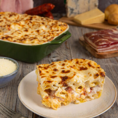 Pasta al forno bianca con crosticina dorata gratinata, servita a fetta con salumi e formaggi sullo sfondo