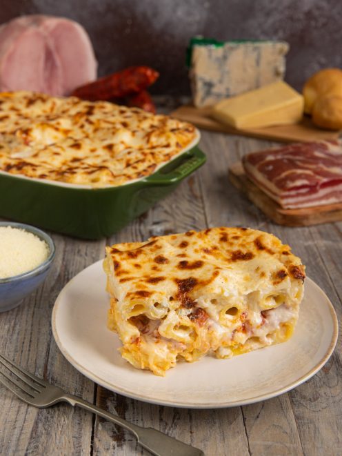 Pasta al forno bianca con crosticina dorata gratinata, servita a fetta con salumi e formaggi sullo sfondo